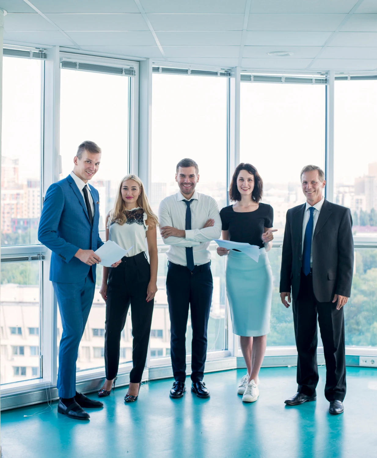 A group of well-dressed professionals standing in a bright, glass-walled office with city views, holding documents and smiling.